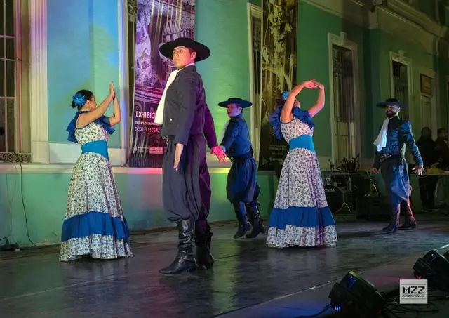 Parejas bailando chacarera con vestidos celestes y trajes de gaucho frente a un edificio histórico